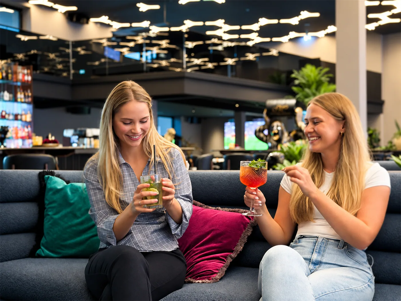 Zwei Gäste sitzen mit Cocktails auf dem Sofa in der Lounge und lachen